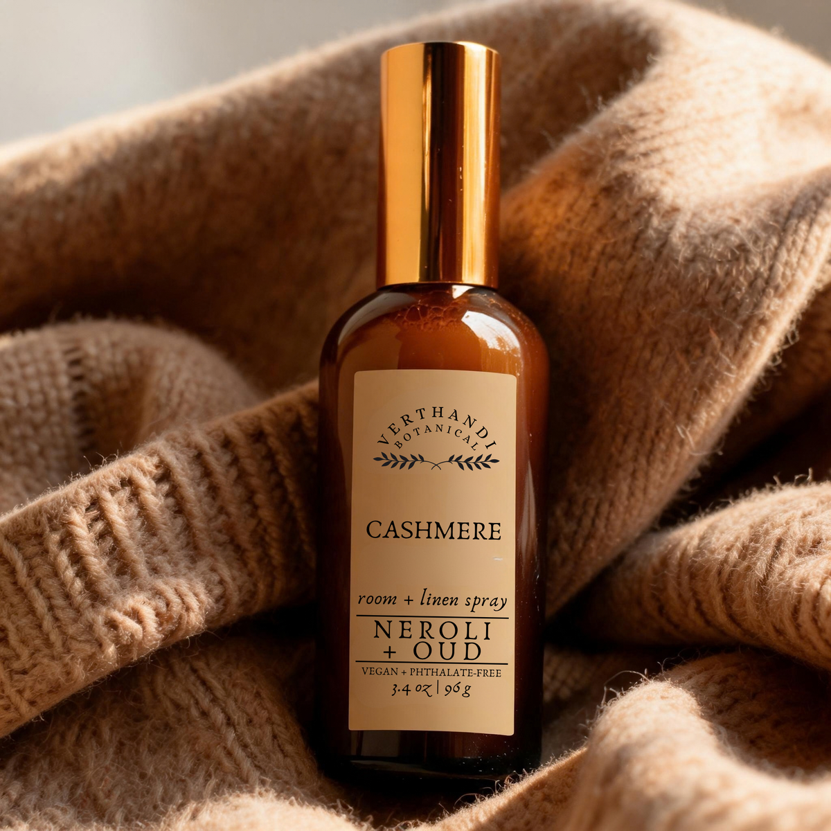 A bottle of Verthandi Botanicals Cashmere Neroli + Oud room and linen spray, featuring an amber glass bottle and a shiny gold cap, resting on a cozy beige knit blanket.